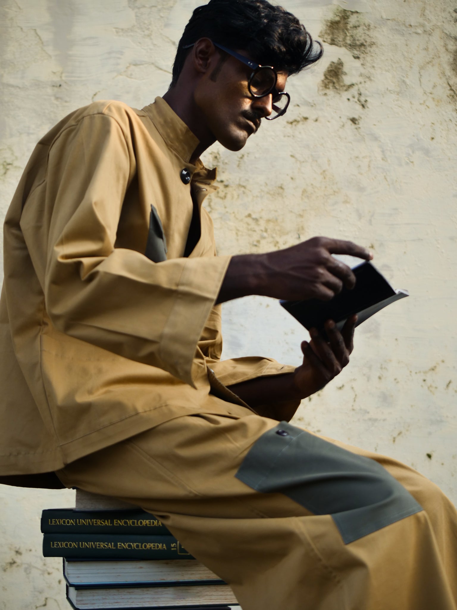 Man in brown outfit with black patches sitting on books, holding a tablet against a textured wall.