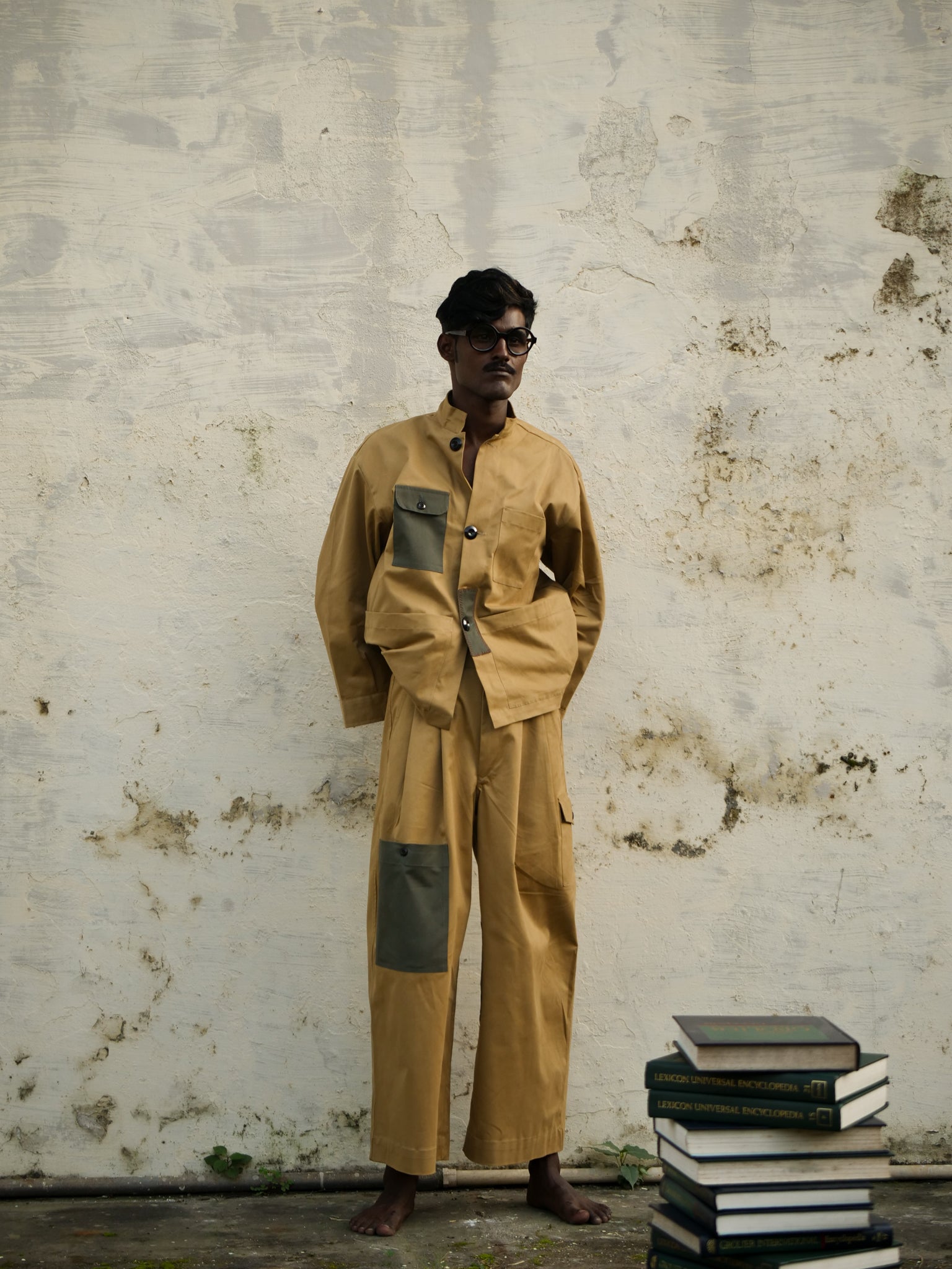 Person wearing a brown jacket and trousers with green patches standing next to a stack of books against a textured wall.