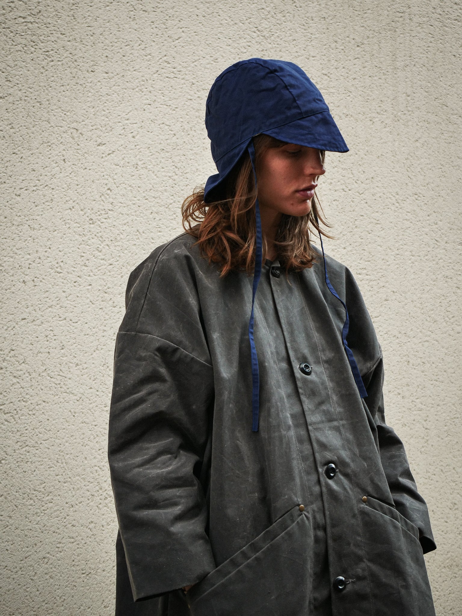 Person wearing a long dark coat and blue bucket hat against a plain background