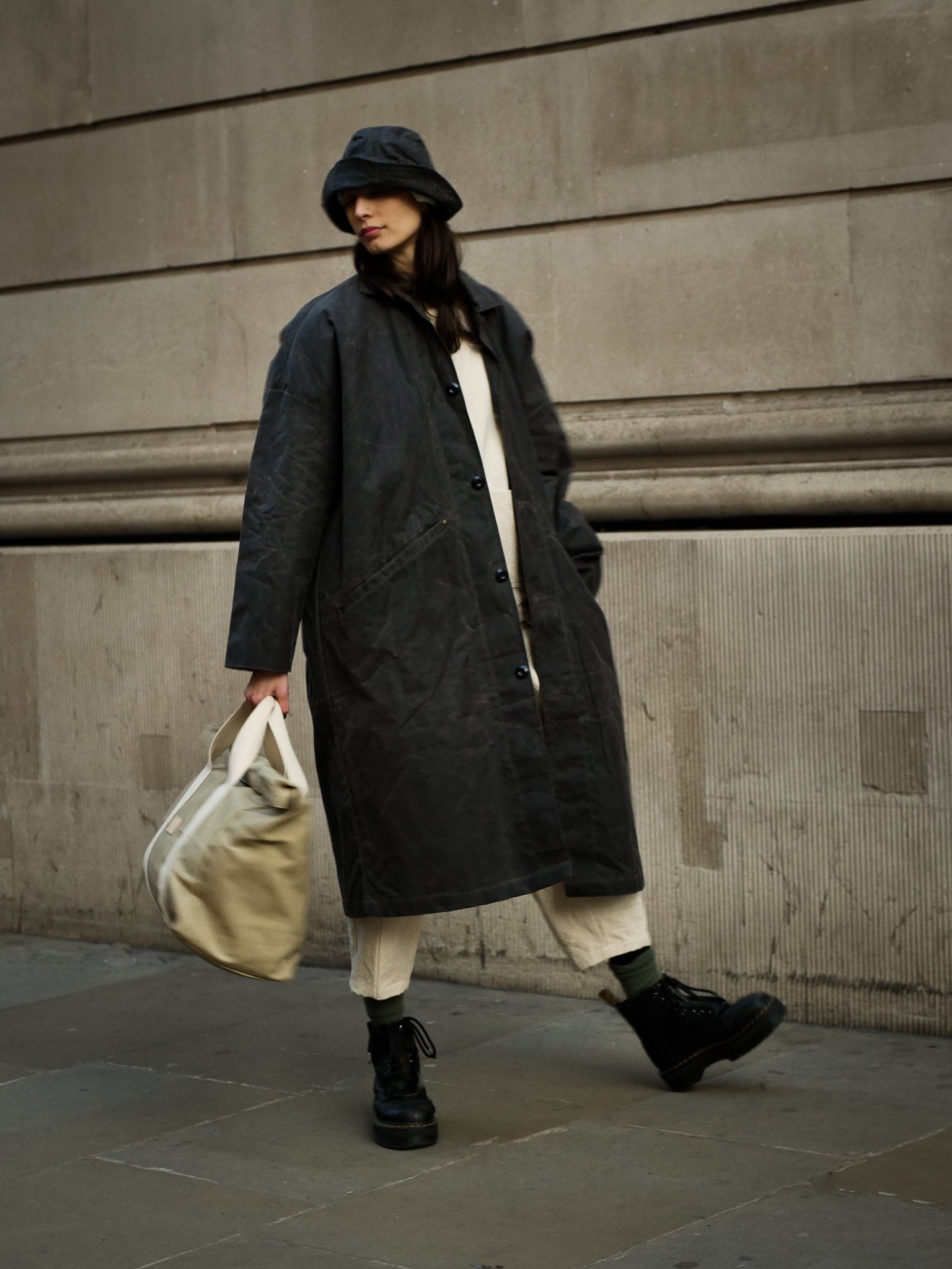 Person wearing a long dark coat and hat, holding a beige bag, against a stone wall.