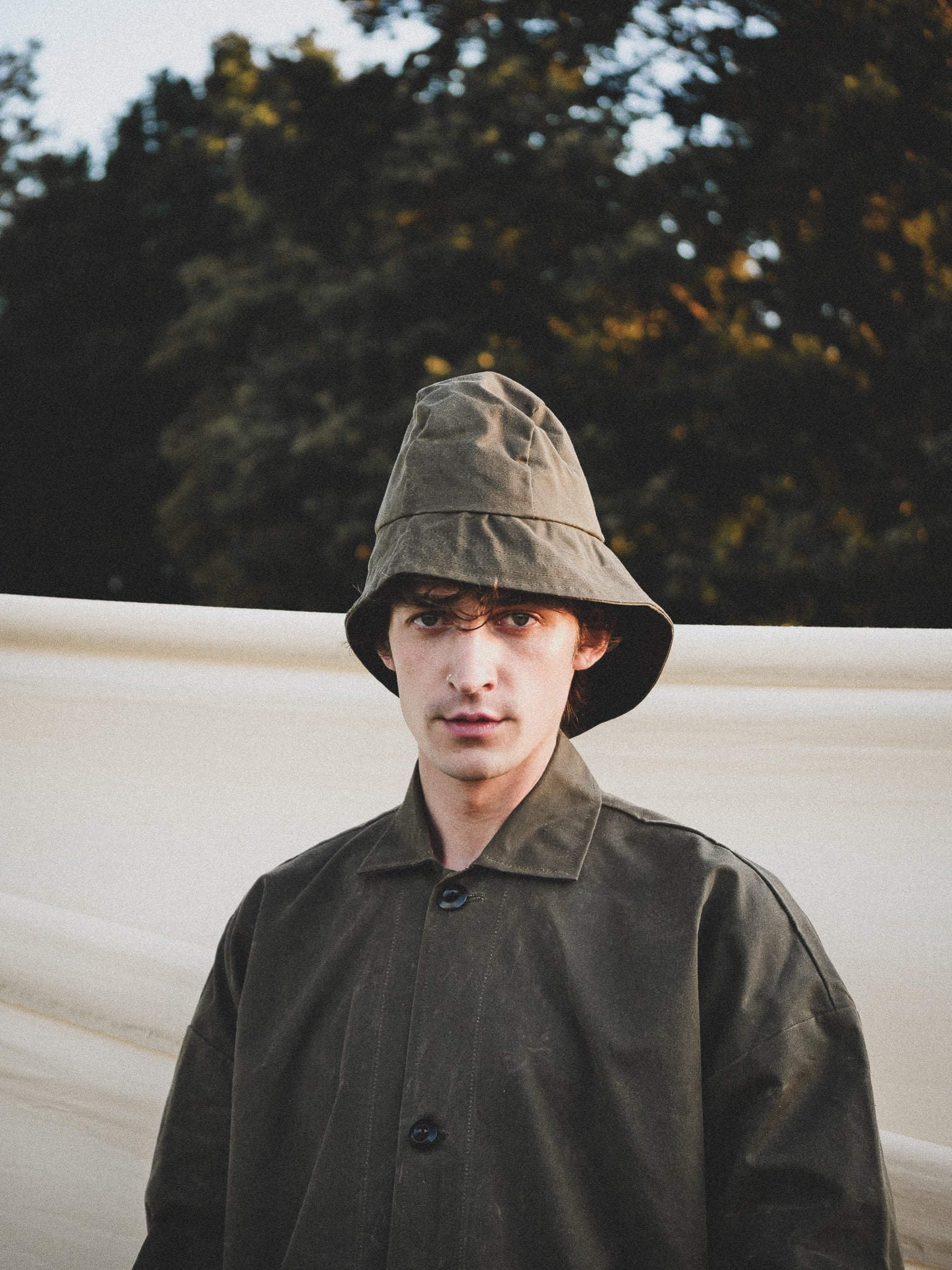 Man wearing a green bucket hat and coat standing outdoors with trees in the background