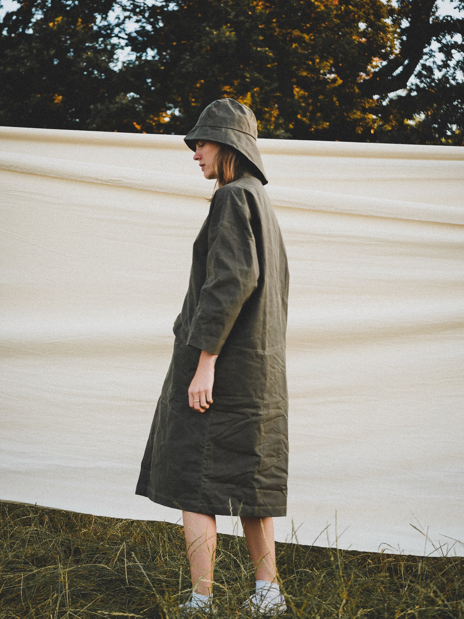 Person wearing a long dark green coat and hat standing in front of a white wall with trees in the background.