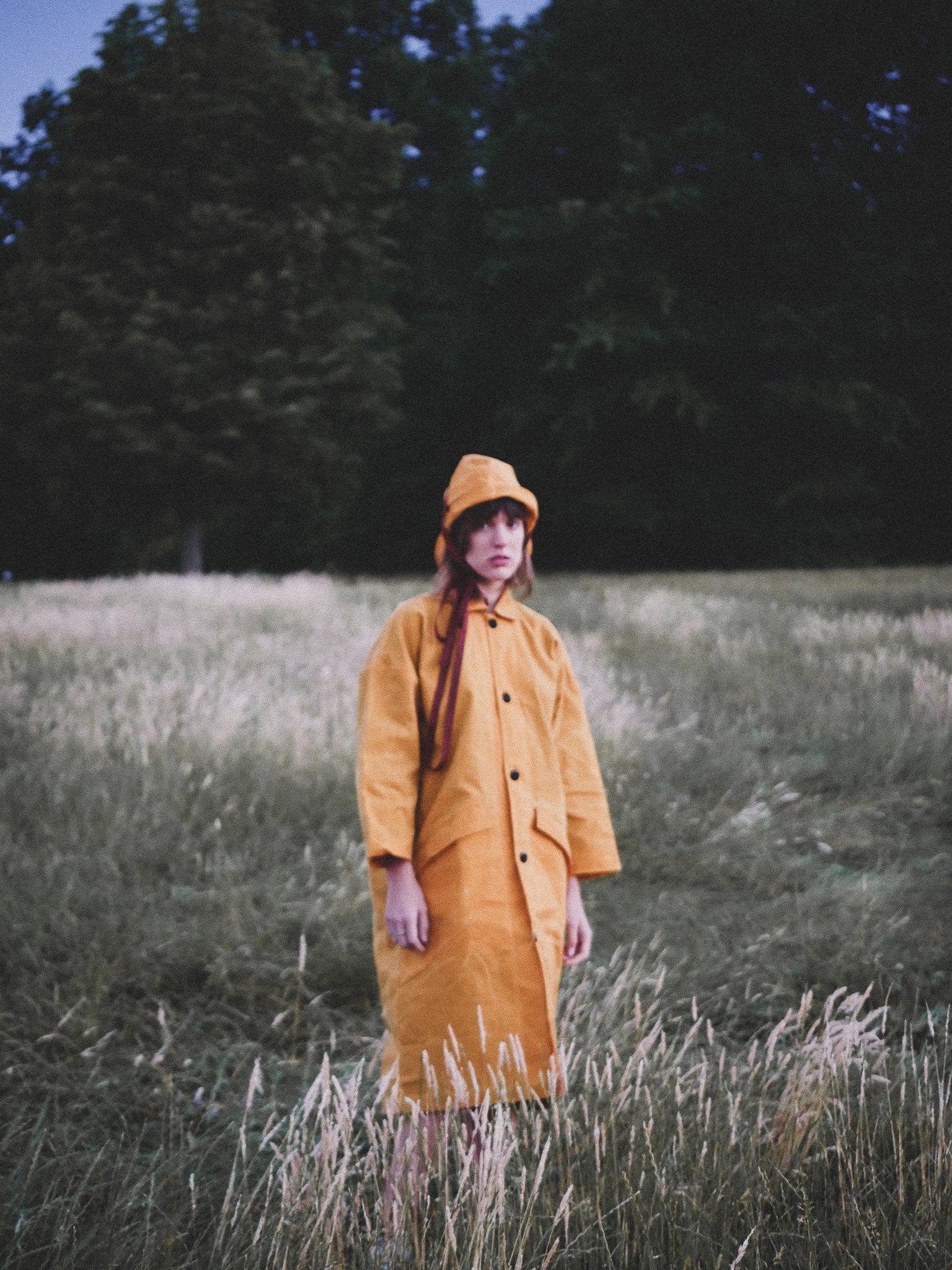 Person wearing a yellow coat and hat standing in a field with trees in the background