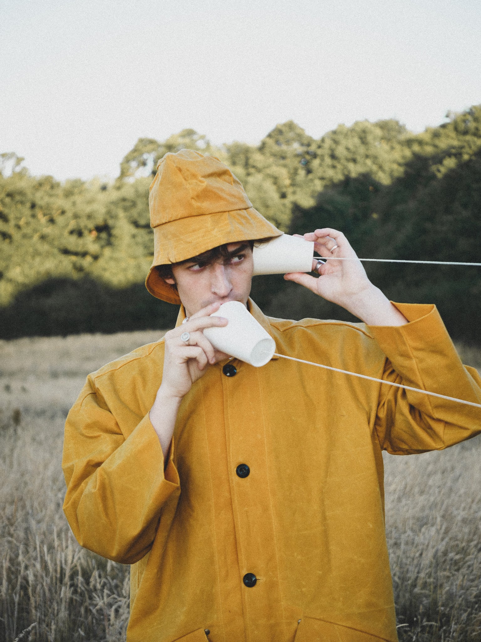 Person in a yellow raincoat and hat drinking from a white cup outdoors.
