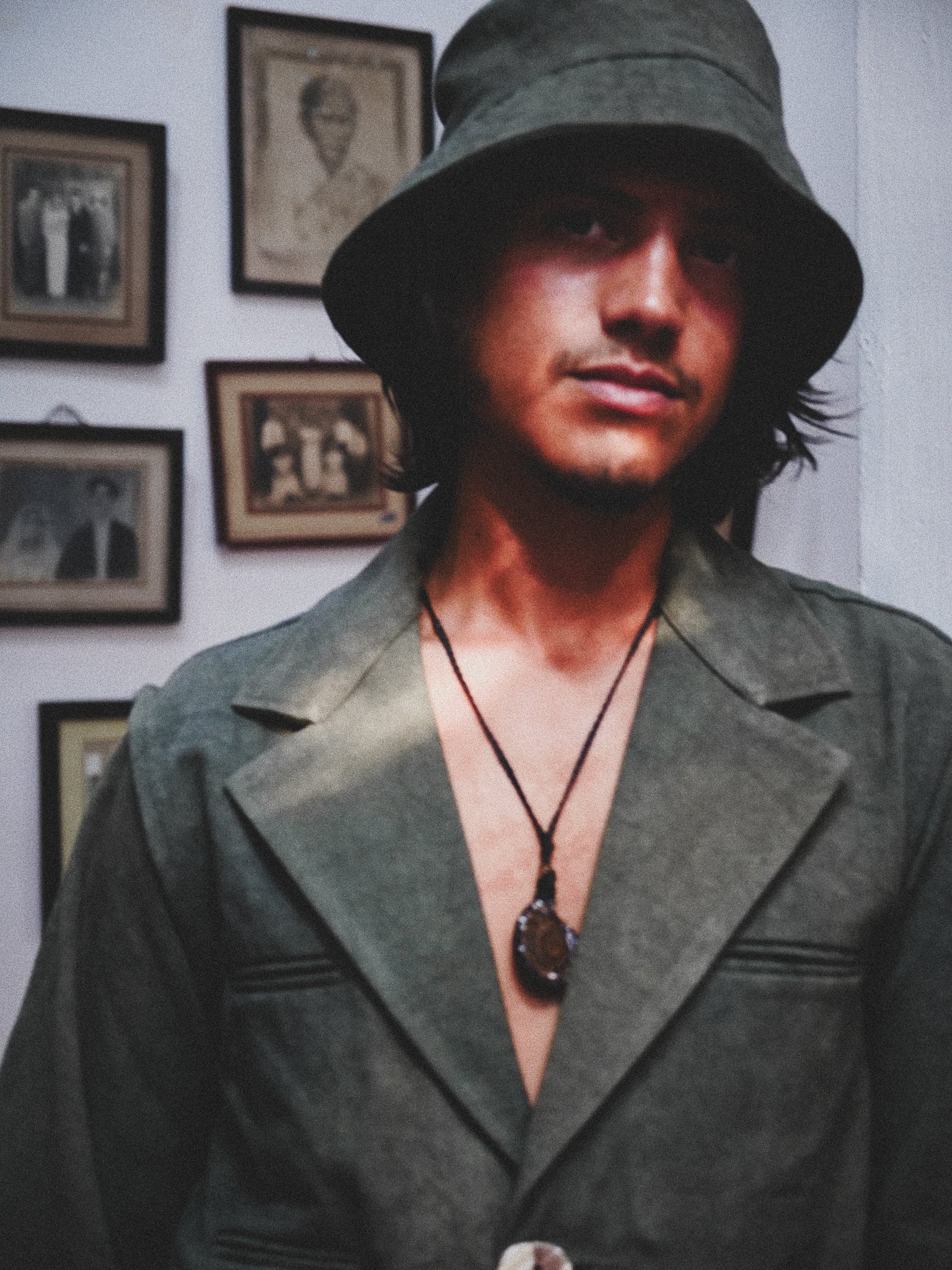 Person wearing a green hat and dark green jacket in front of framed pictures on a wall.