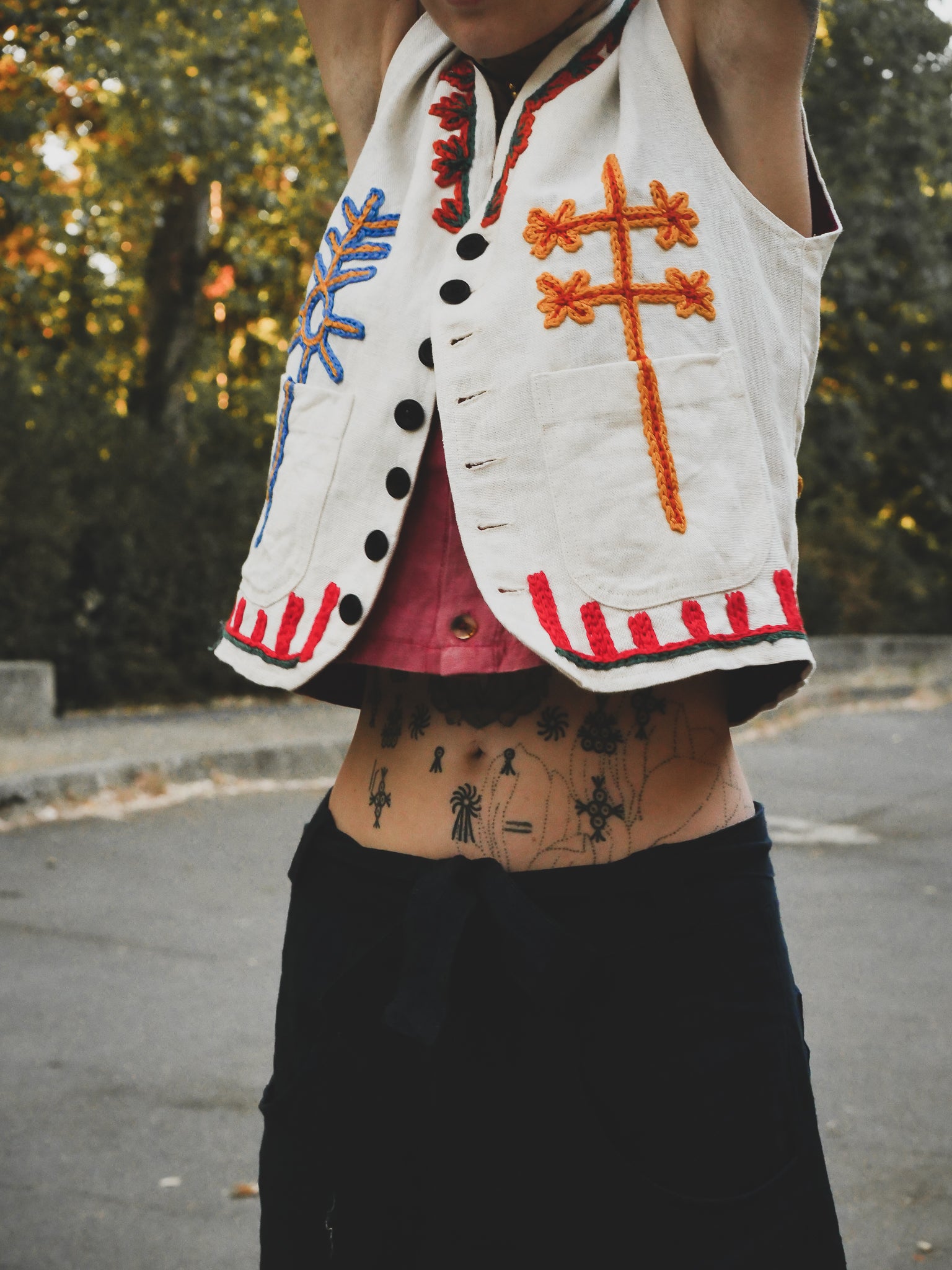 Person wearing a white vest with colorful hand embroidery, hanging outdoors.
