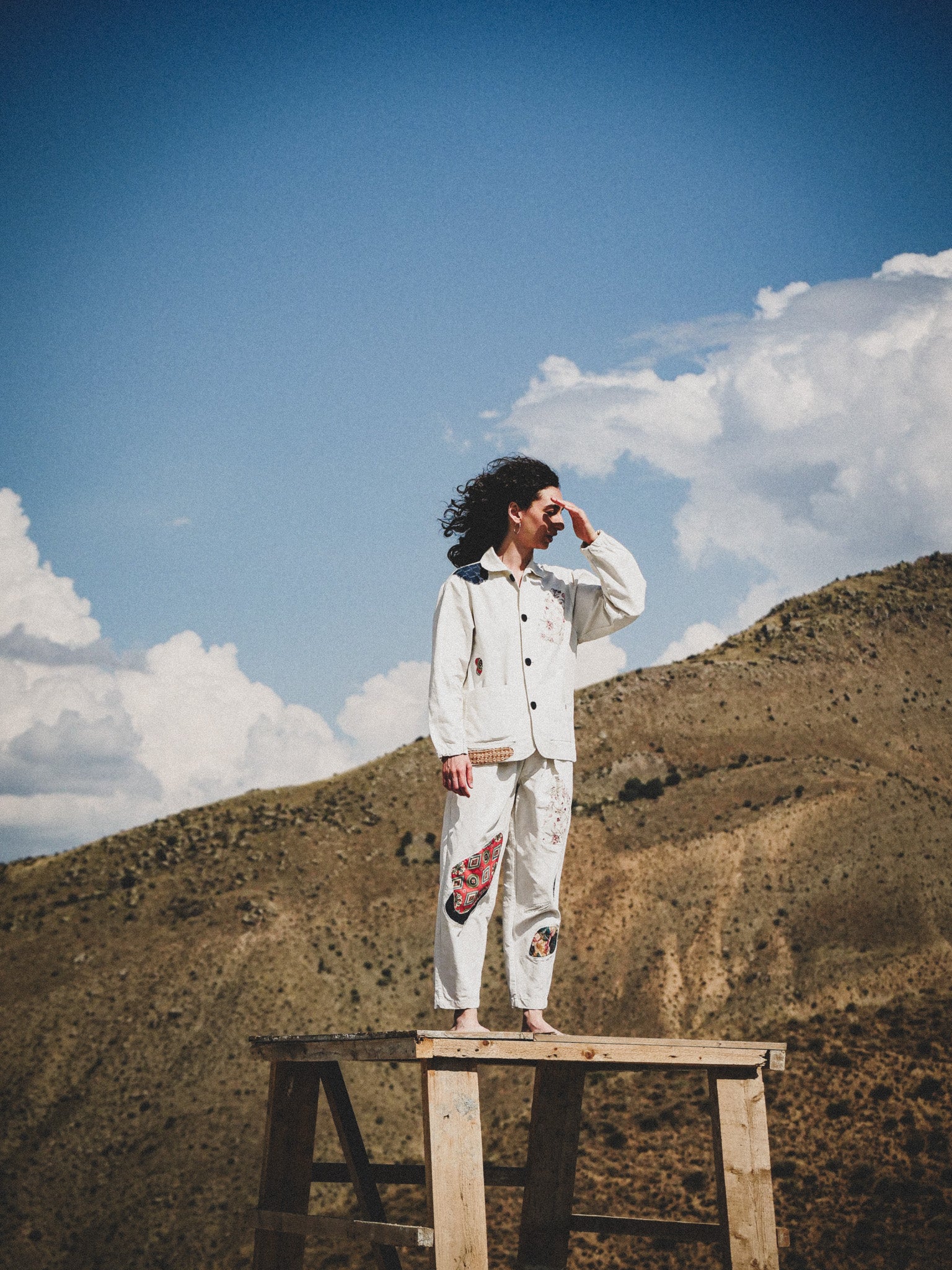 Person in a white outfit with patterned patchwork standing on a wooden platform against a mountainous landscape.