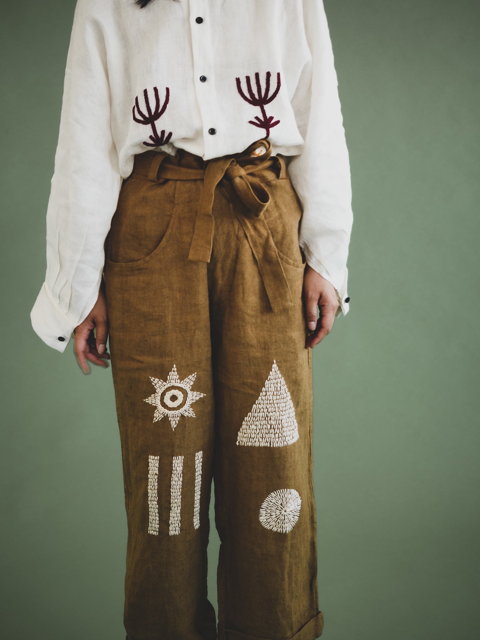 Person wearing a white shirt with embroidered designs and brown pants with geometric patterns on a green background