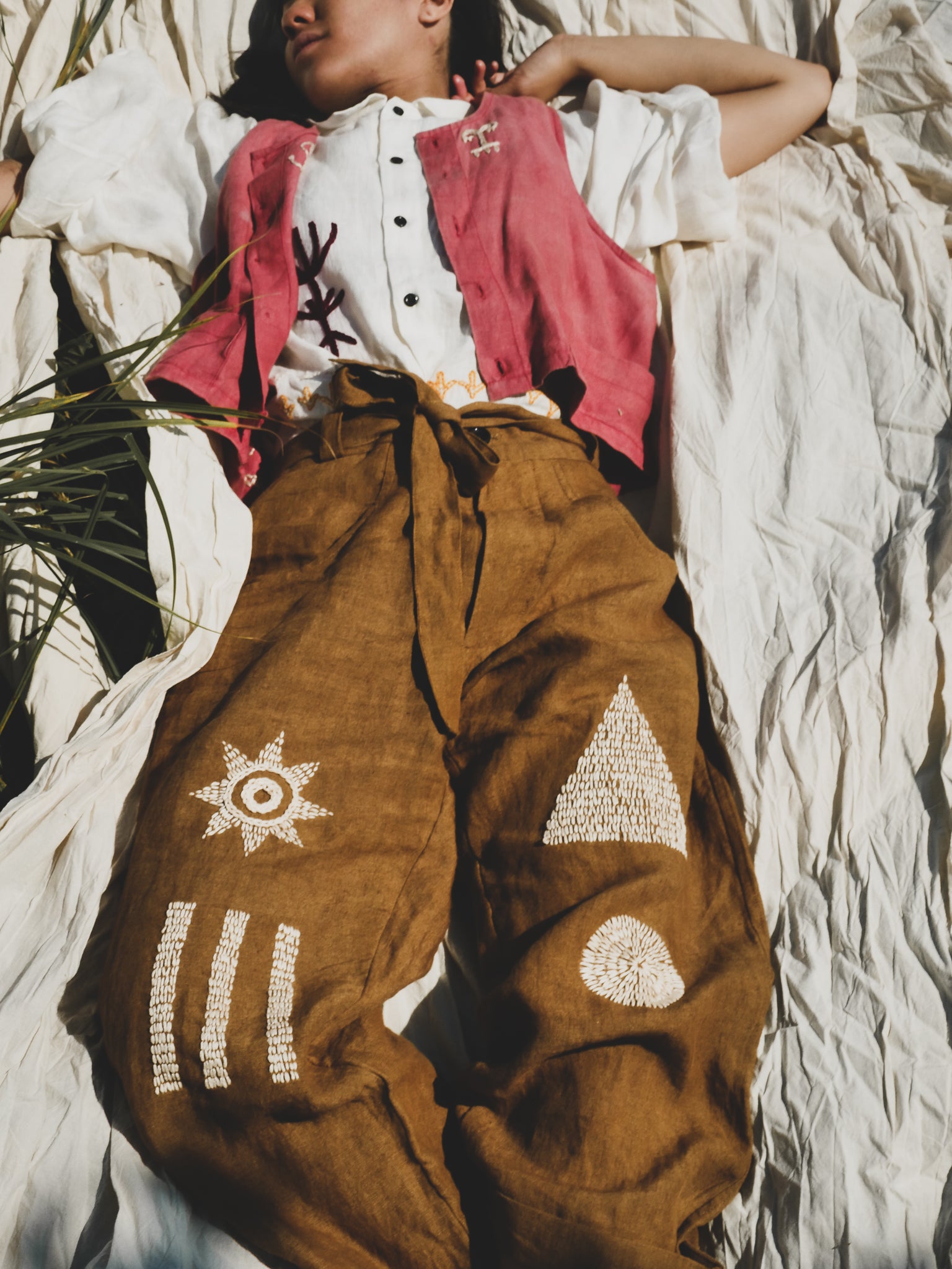 Person wearing a white shirt, red jacket, and brown pants with hand embroidered patterns, lying on a textured surface.