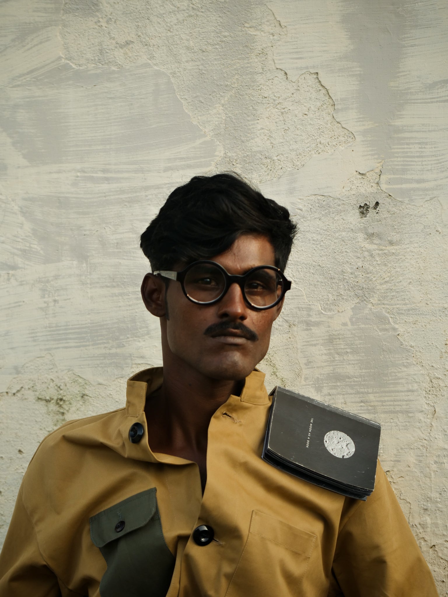 Person wearing a brown jacket with a book on his shoulder against a textured wall.