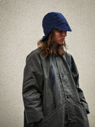 Person wearing a long dark coat and blue bucket hat against a plain background