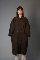 Person wearing a long brown coat against a gray background