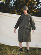 Person wearing a long green coat and hat standing outdoors with trees in the background