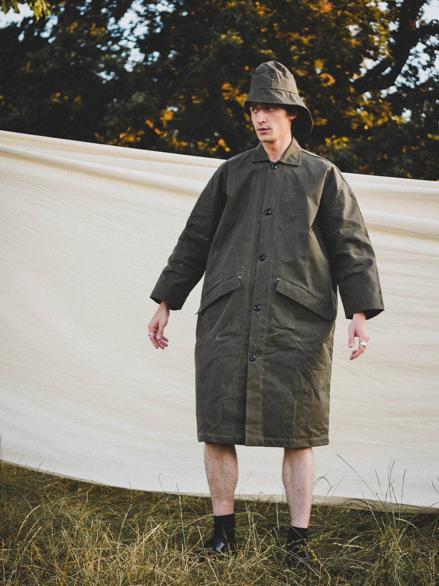 Person wearing a long green coat and hat standing outdoors with trees in the background