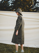 Person wearing a long dark green coat and hat standing in front of a white wall with trees in the background.