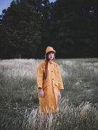 Person wearing a yellow coat and hat standing in a field with trees in the background