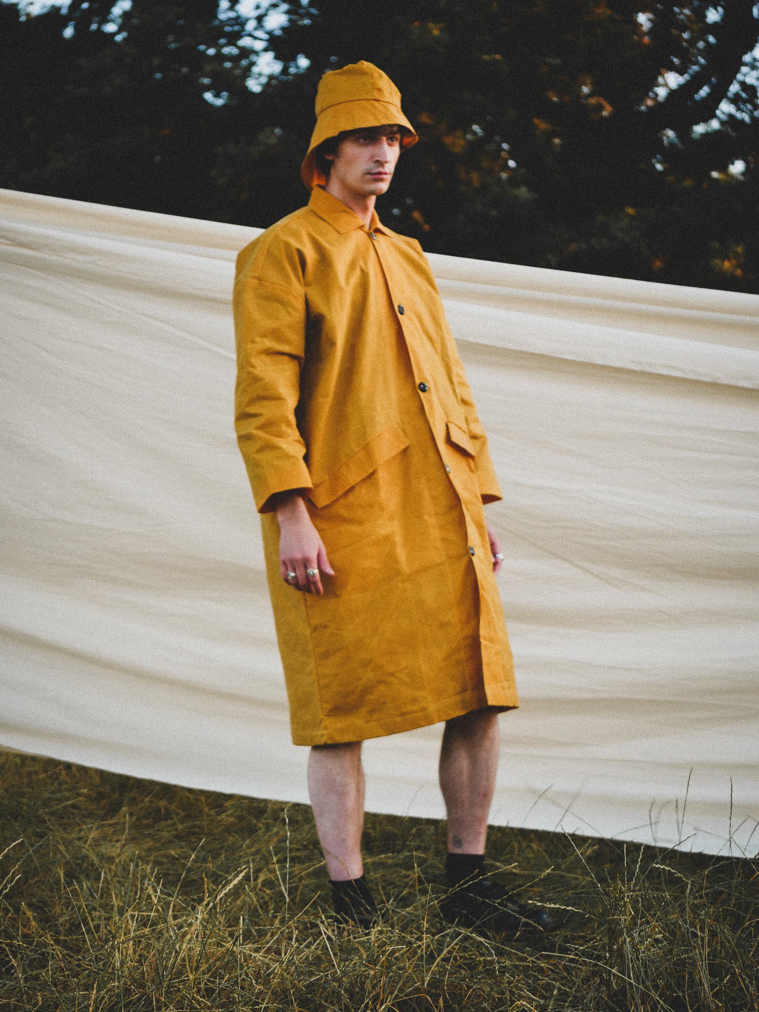 Person wearing a mustard yellow raincoat and hat standing in front of a white fabric backdrop.