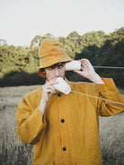 Person in a yellow raincoat and hat drinking from a white cup outdoors.