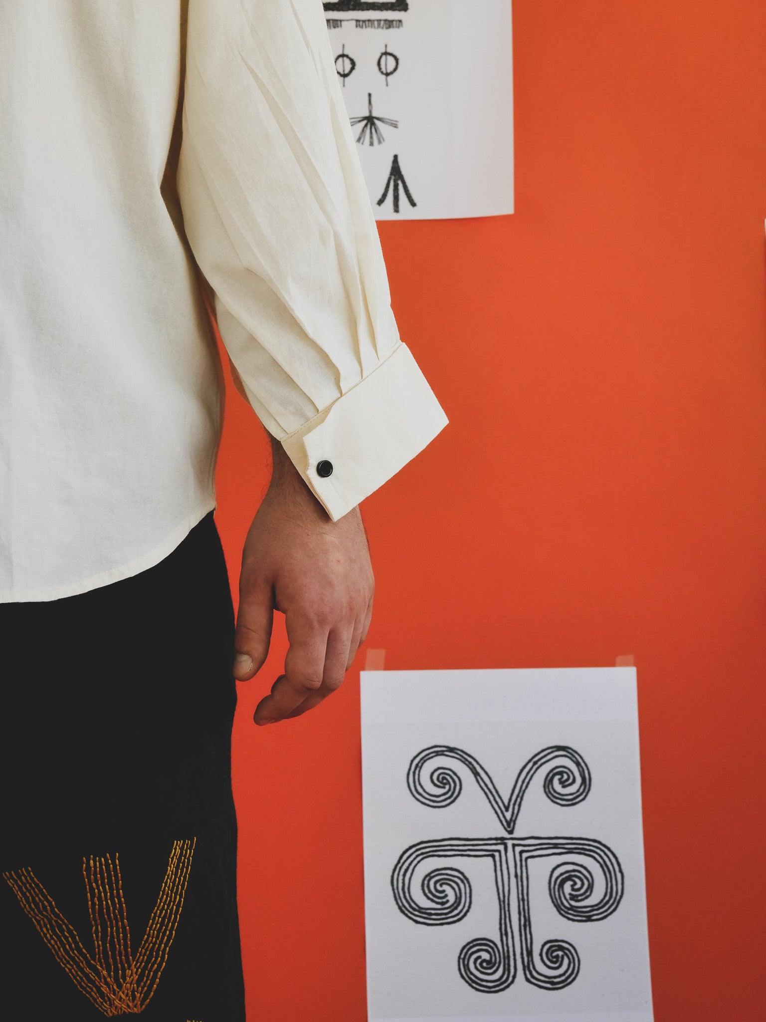 Person wearing an off-white shirt with cuffs against an orange wall with black and white designs.