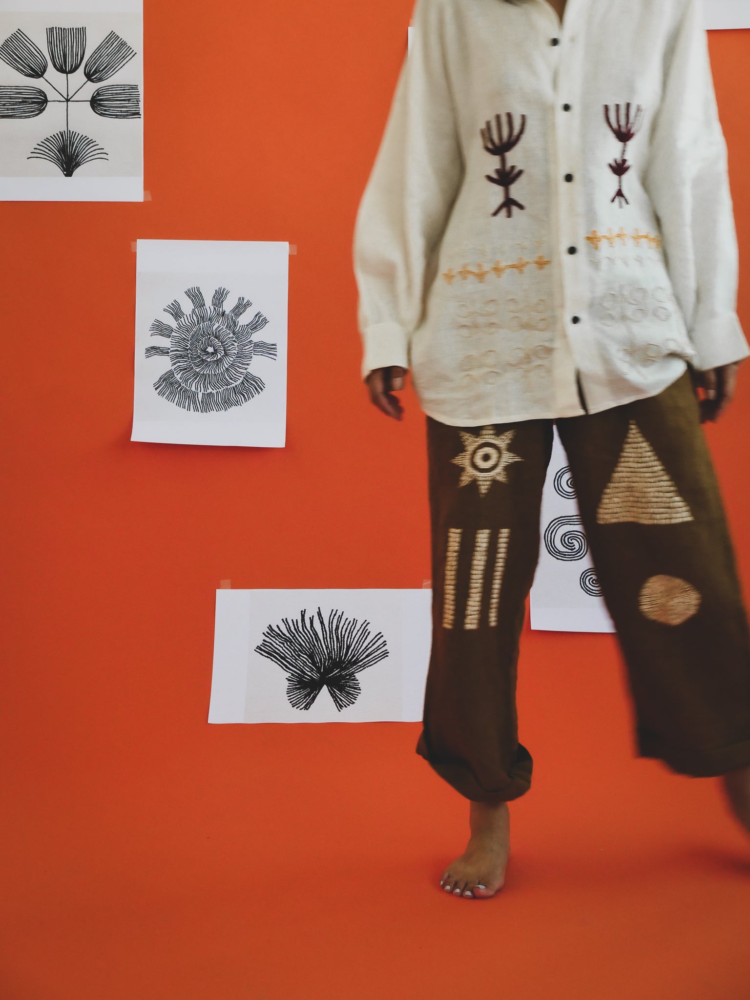 Person wearing a white shirt with embroidered designs on an orange wall with black and white prints.