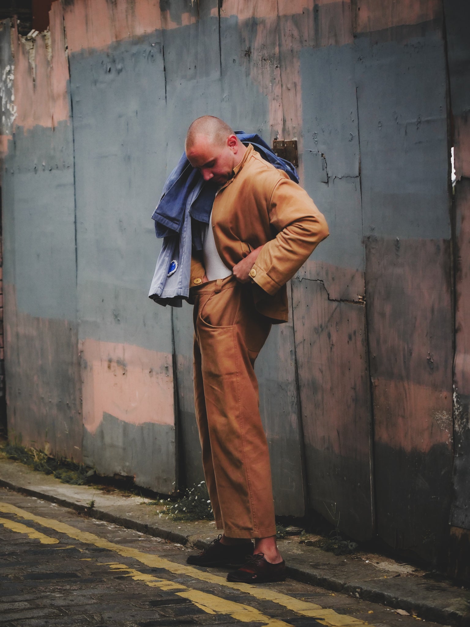 Man in a brown coat leaning against a textured wall.