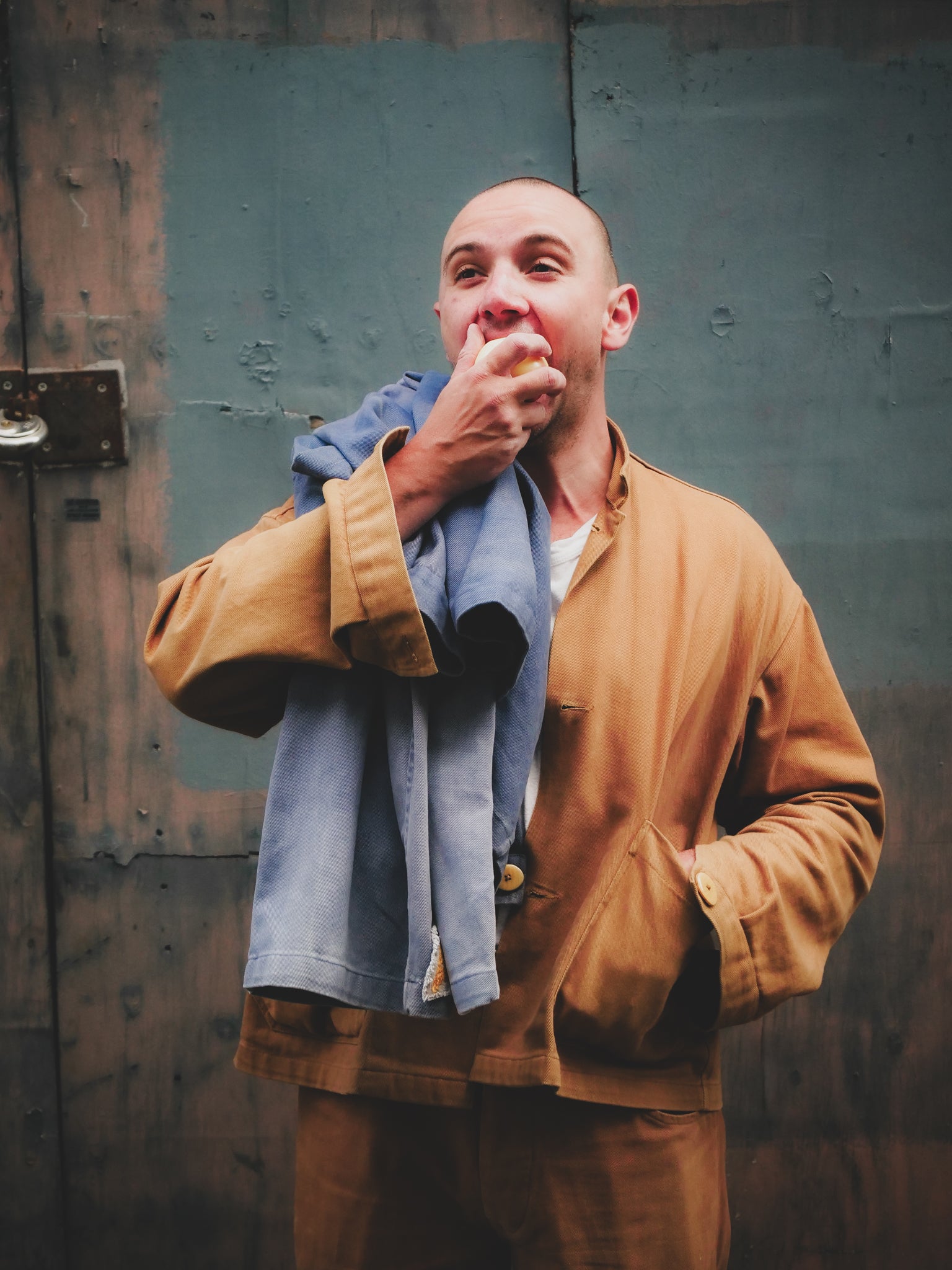 Man in a tan jacket eating an orange against a textured wall.