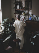Person in a white suit standing in a room with plants and shelves.
