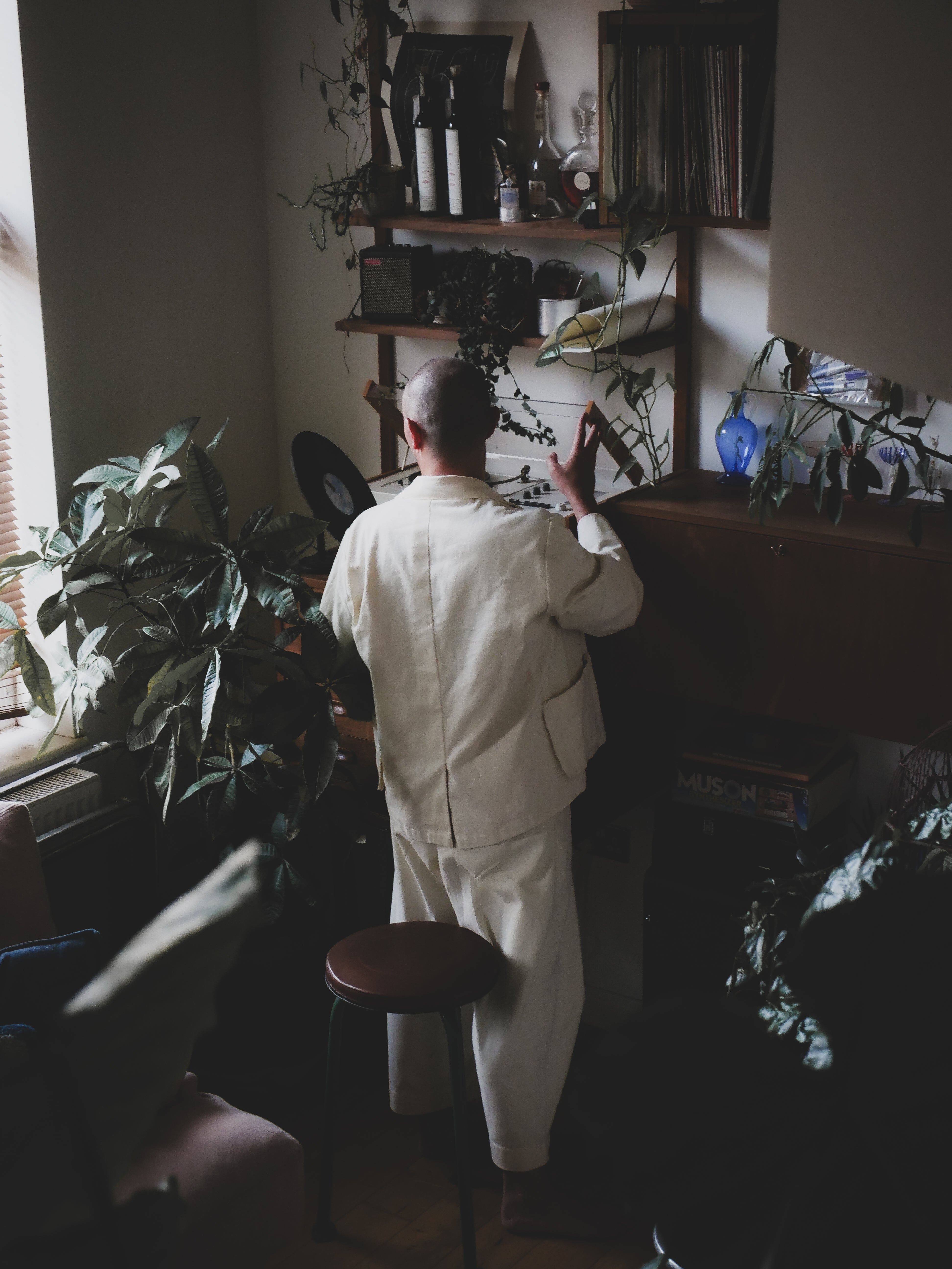 Person in a white suit standing in a room with plants and shelves.