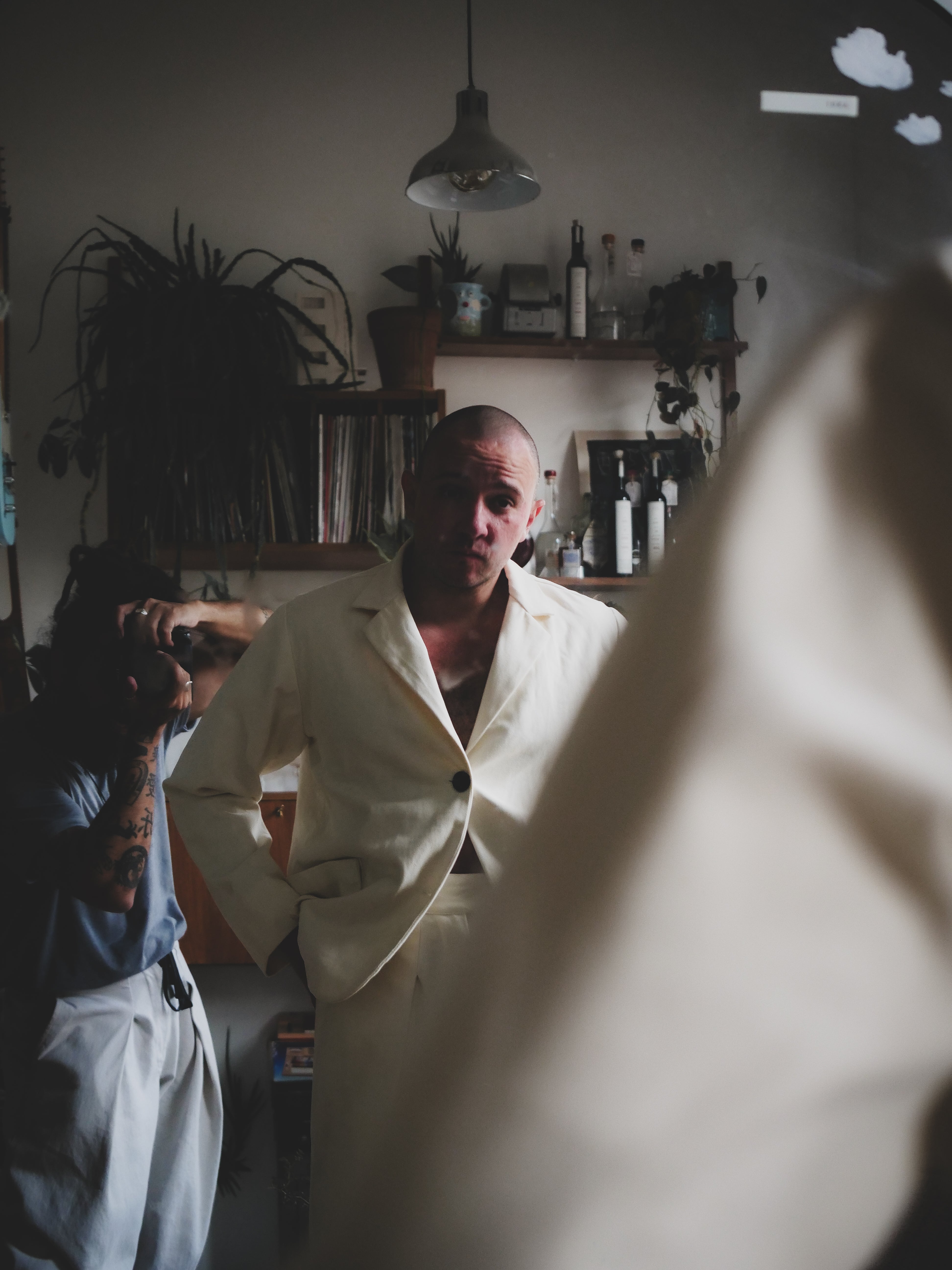 Man in a beige suit standing in a room with plants and shelves.