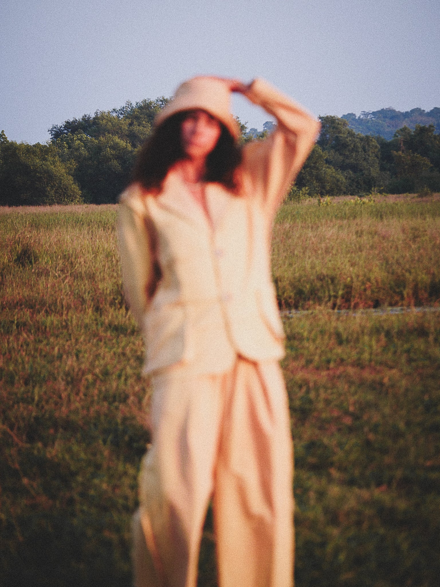 Person in a pale mustard outfit standing in a field with trees in the background