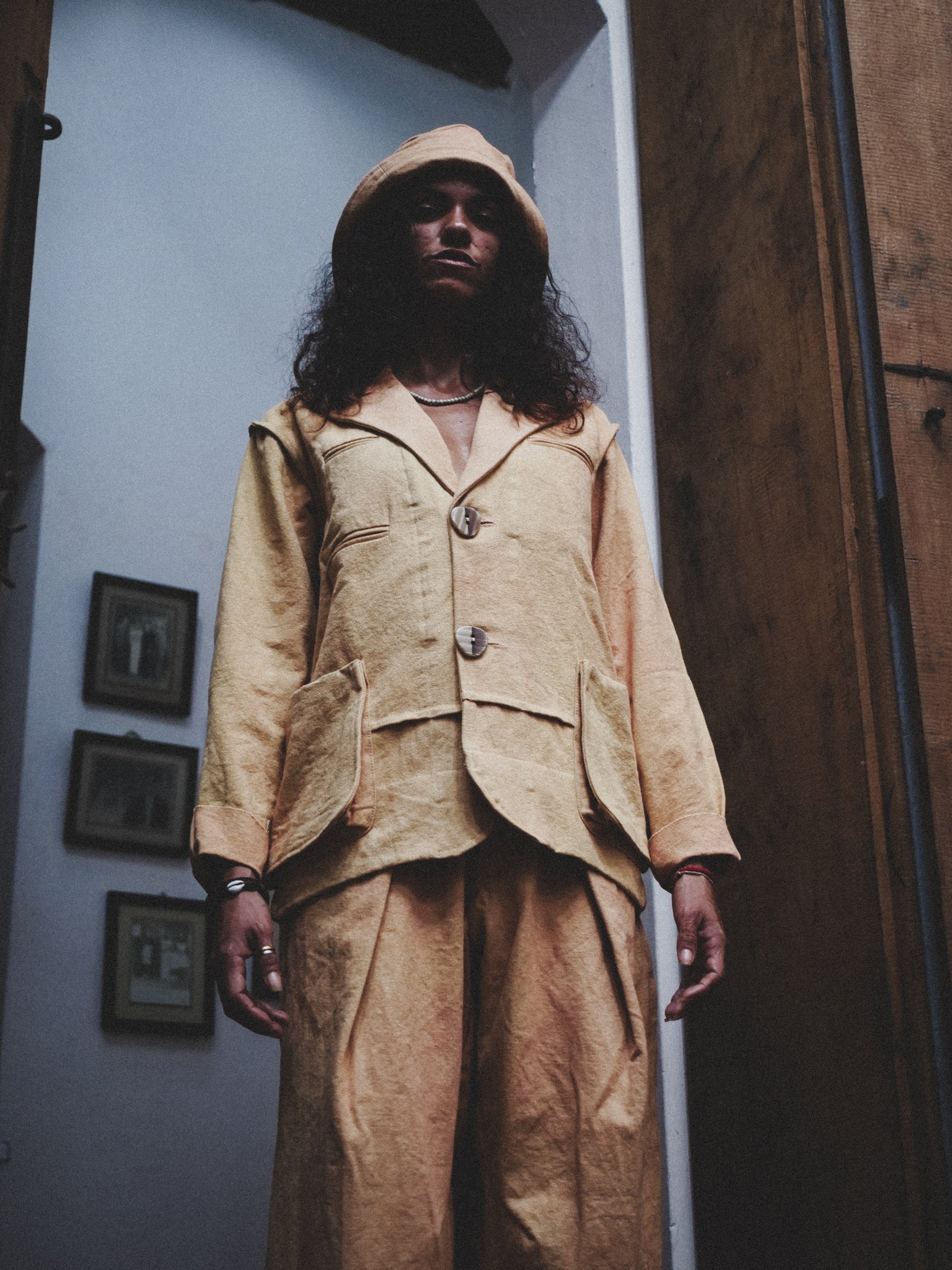 Person wearing a pale mustard suit standing in a room with framed pictures on the wall.