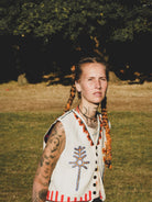 Person with braided hair and tattoos wearing a vest with a hand embroidered design, standing in a grassy field.