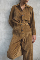 Person wearing a brown corduroy jumpsuit against a gray concrete wall.