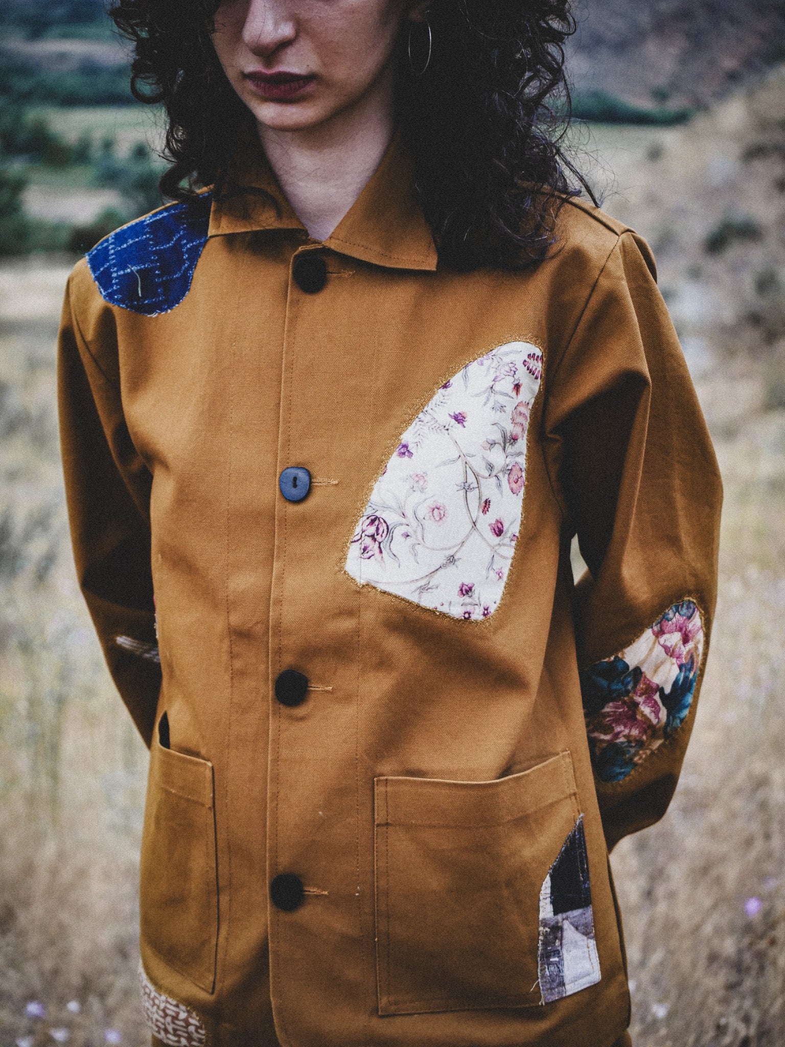 Person wearing a brown jacket with patchwork sleeves and chest, standing in a natural setting.