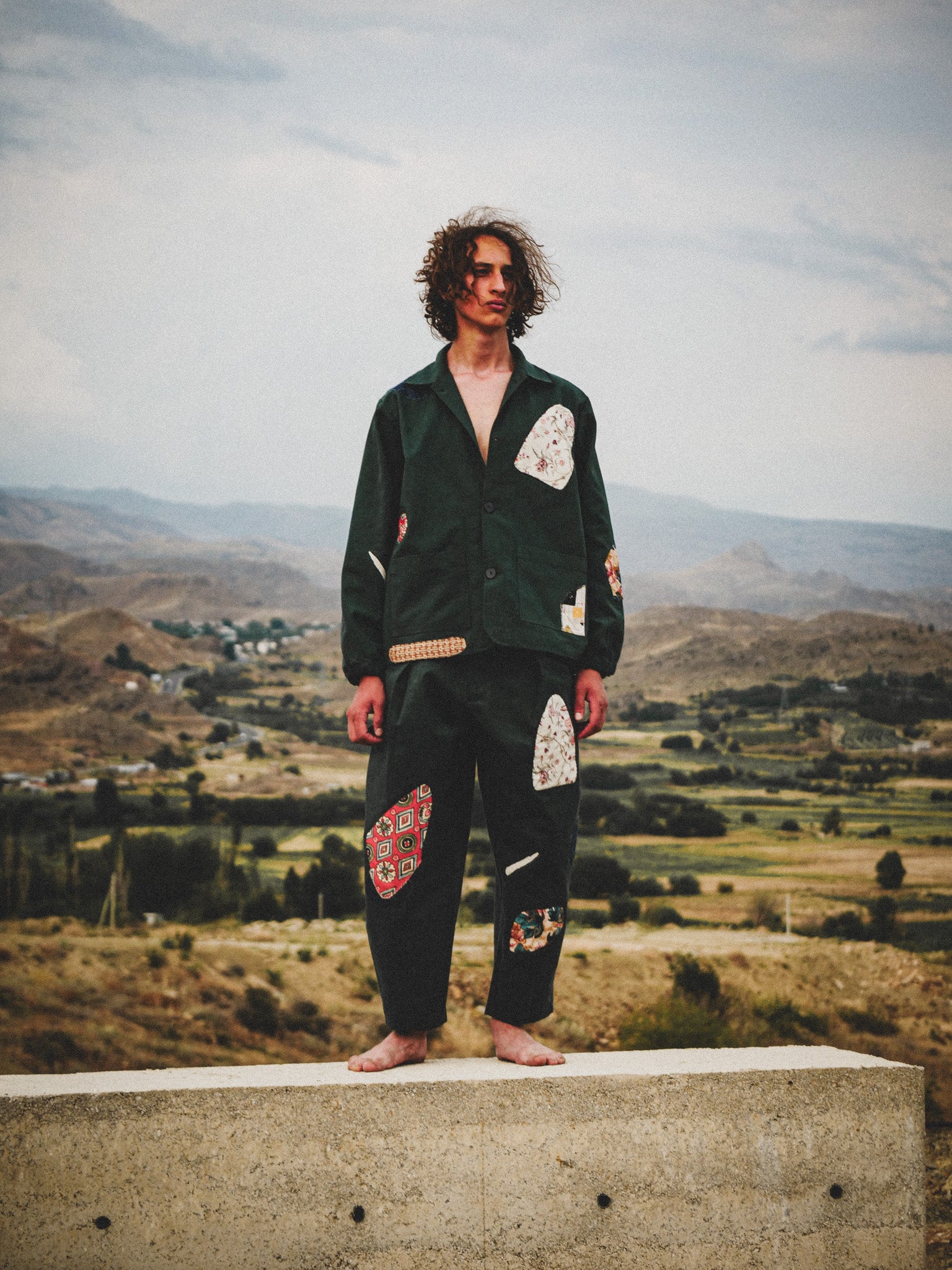 Person wearing a green jacket and patterned pants standing on a ledge with a scenic landscape in the background