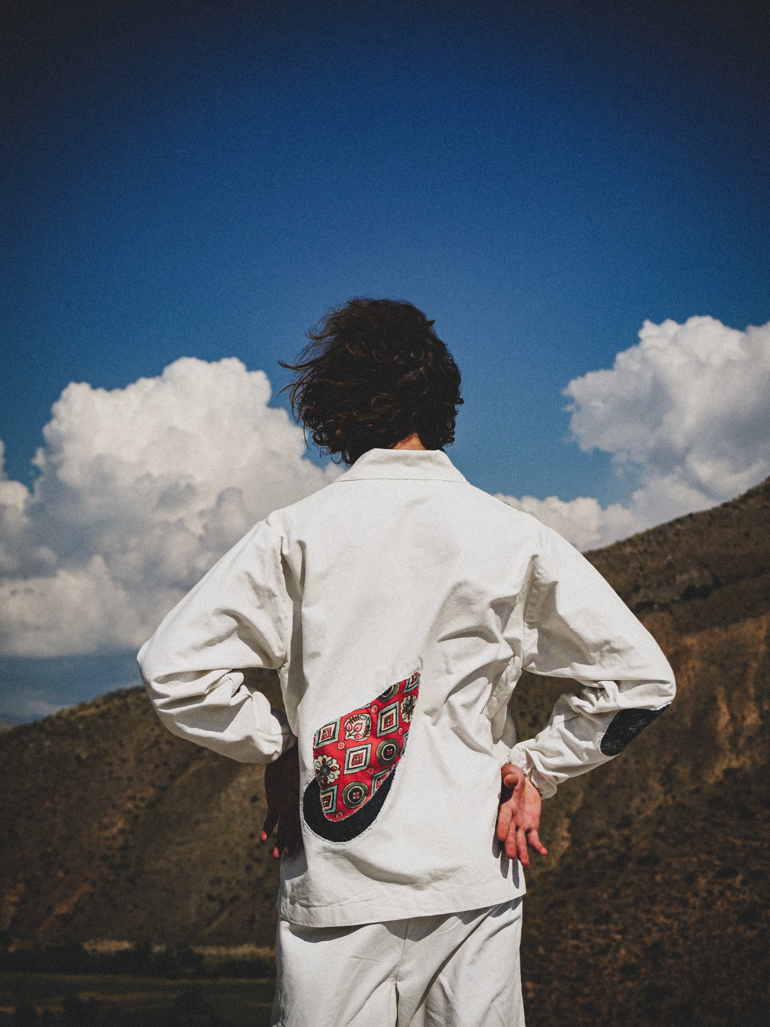 Person wearing a white jacket with a colorful pattern on the back, standing against a mountainous landscape with clouds.