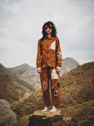Person wearing a brown outfit with patches standing on a rock in a mountainous landscape