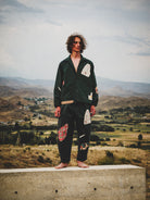 Person wearing a green jacket and patterned pants standing on a ledge with a scenic landscape in the background