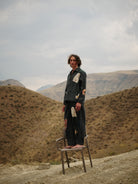 Person in green trousers with patterned patchwork standing on a chair in a desert landscape with mountains in the background
