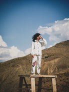 Person in a white outfit with patterned patchwork standing on a wooden platform against a mountainous landscape.