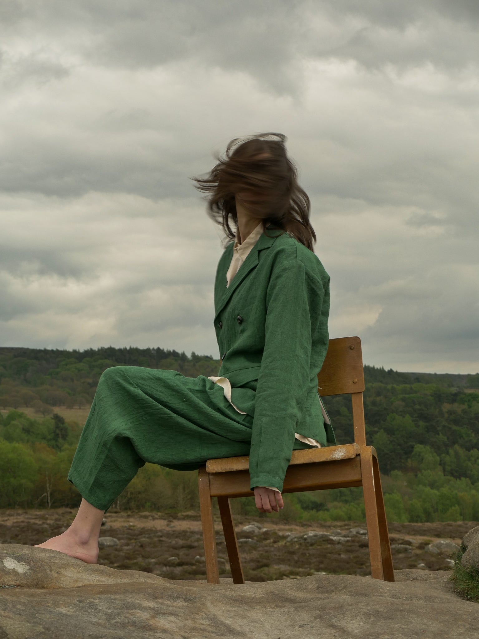 Person in a green outfit sitting on a wooden chair against a backdrop of a cloudy sky and forest.