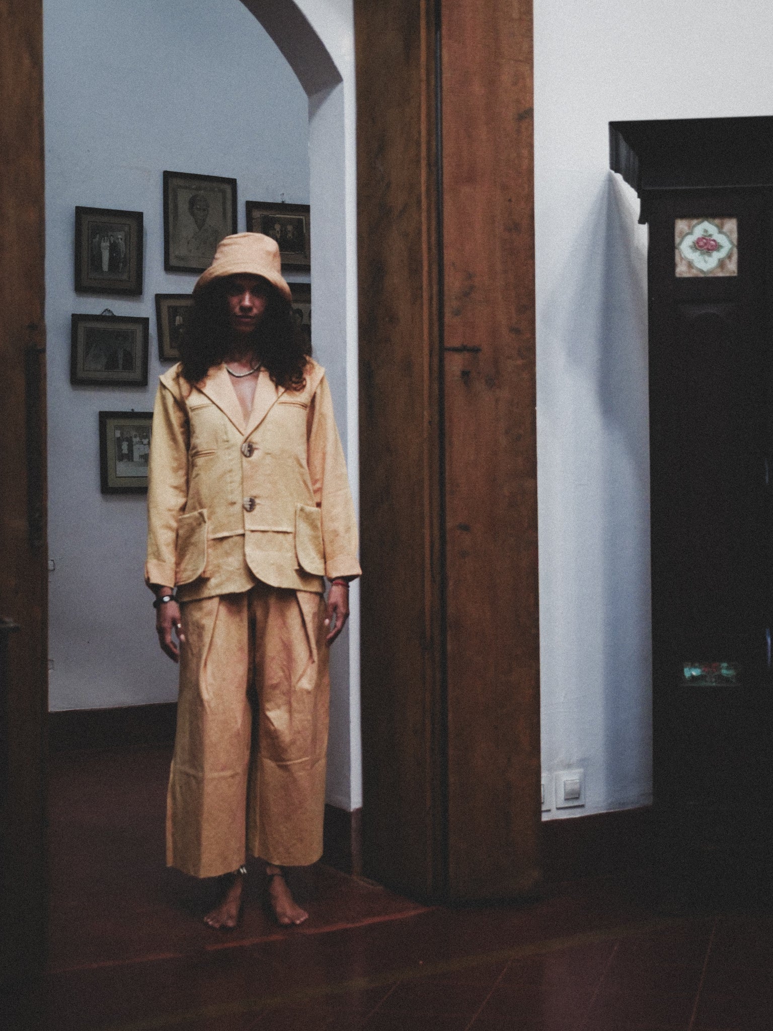 Person wearing a pale mustard suit standing in a room with wooden doors and framed pictures on the wall.