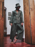 Person wearing a dark green suit and hat in a room with wooden floors and framed pictures on the wall.