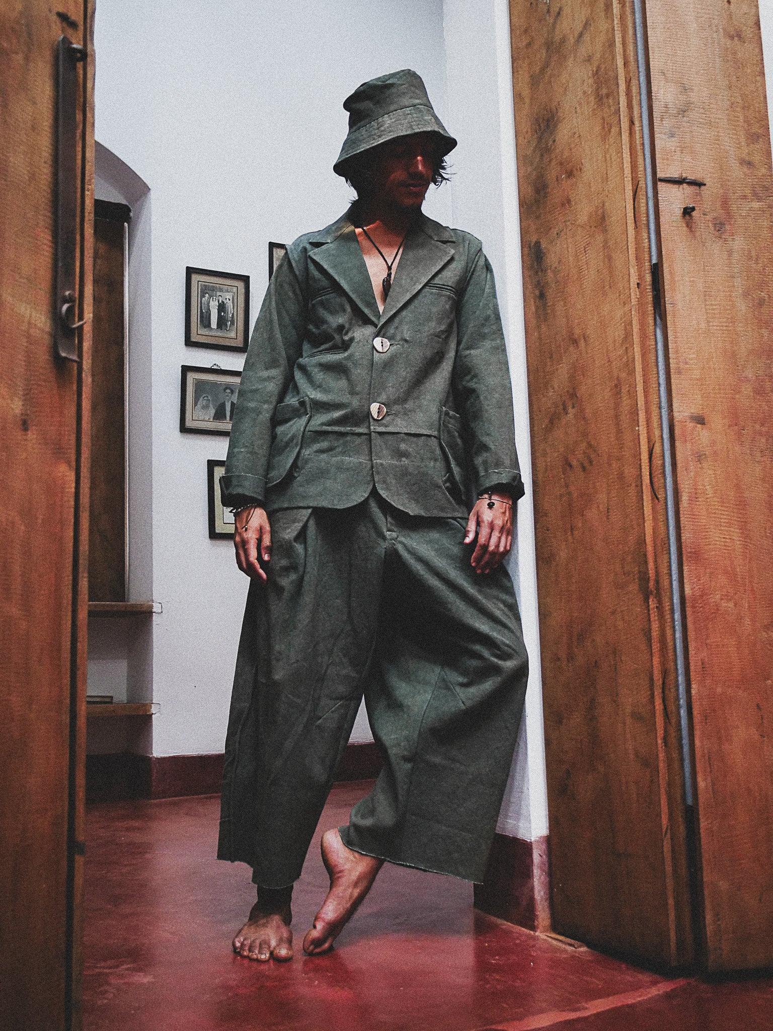 Person wearing a dark green suit and hat in a room with wooden floors and framed pictures on the wall.