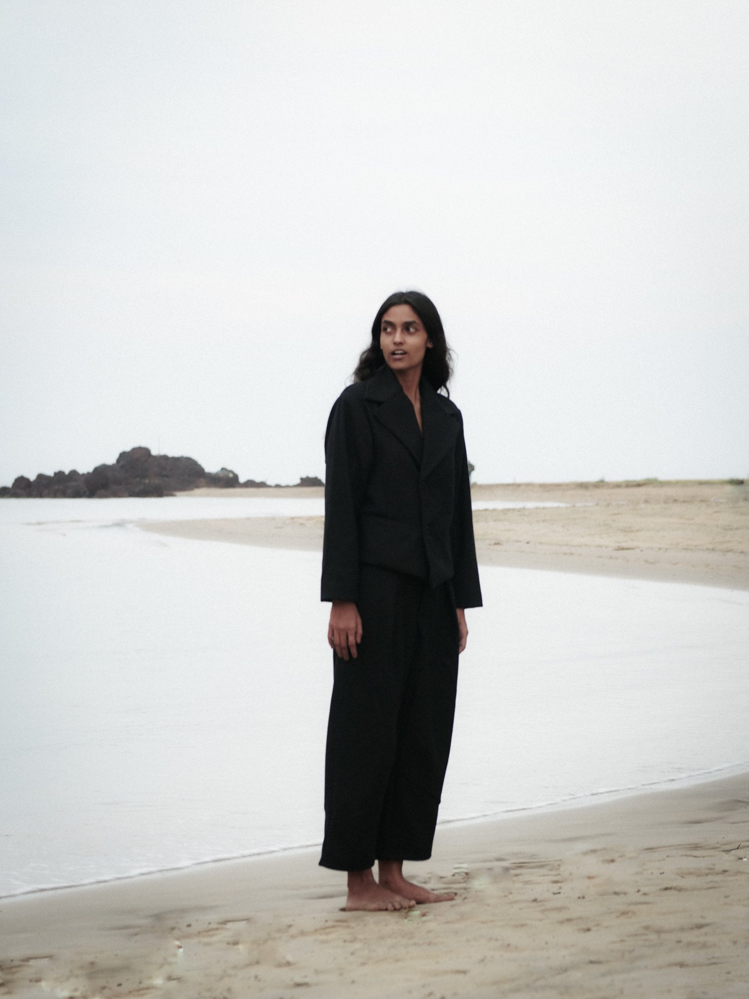 Person wearing a black outfit standing on a beach with a minimalistic background