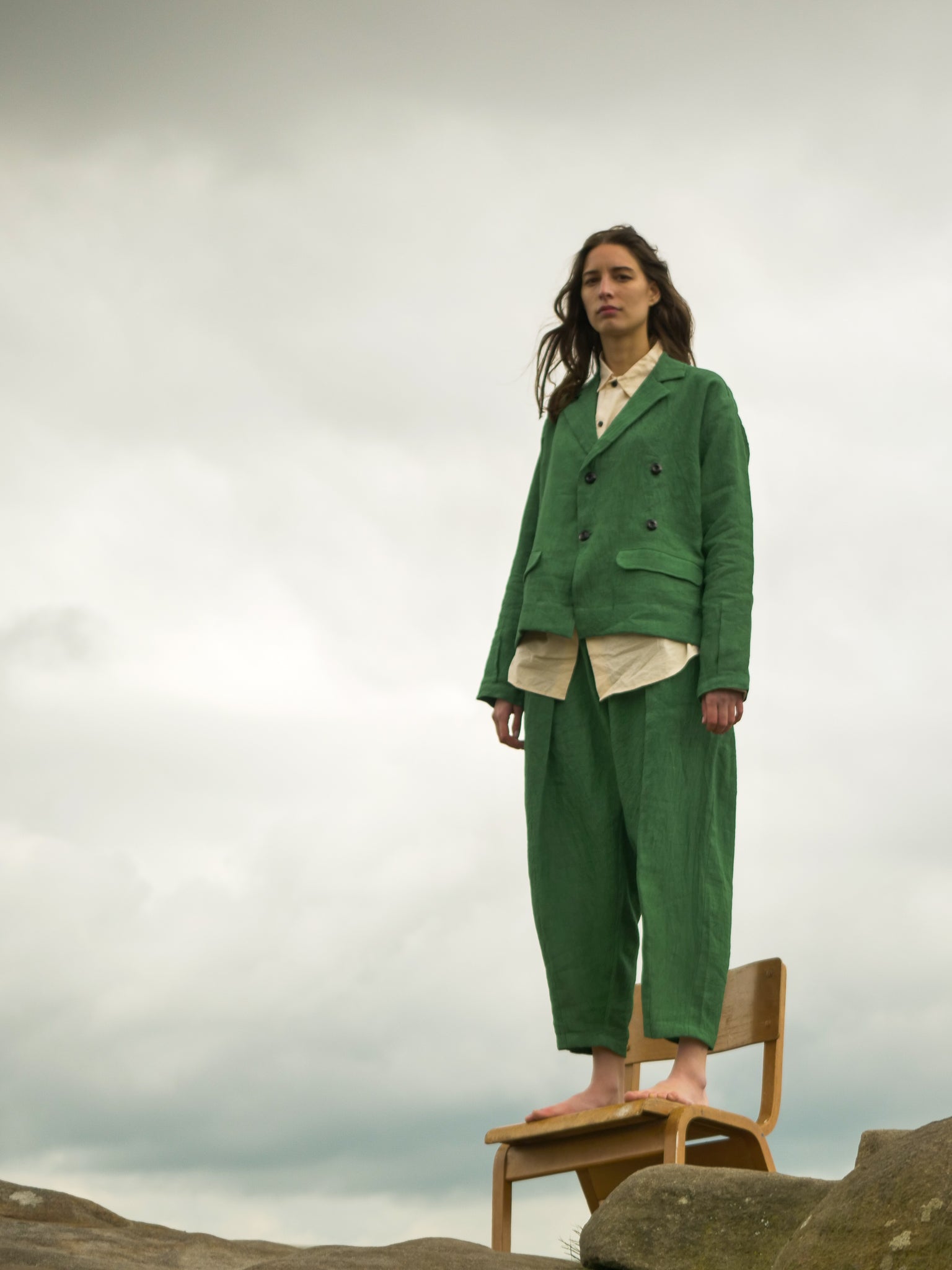Person wearing a green suit standing on a rock with a chair and cloudy sky in the background