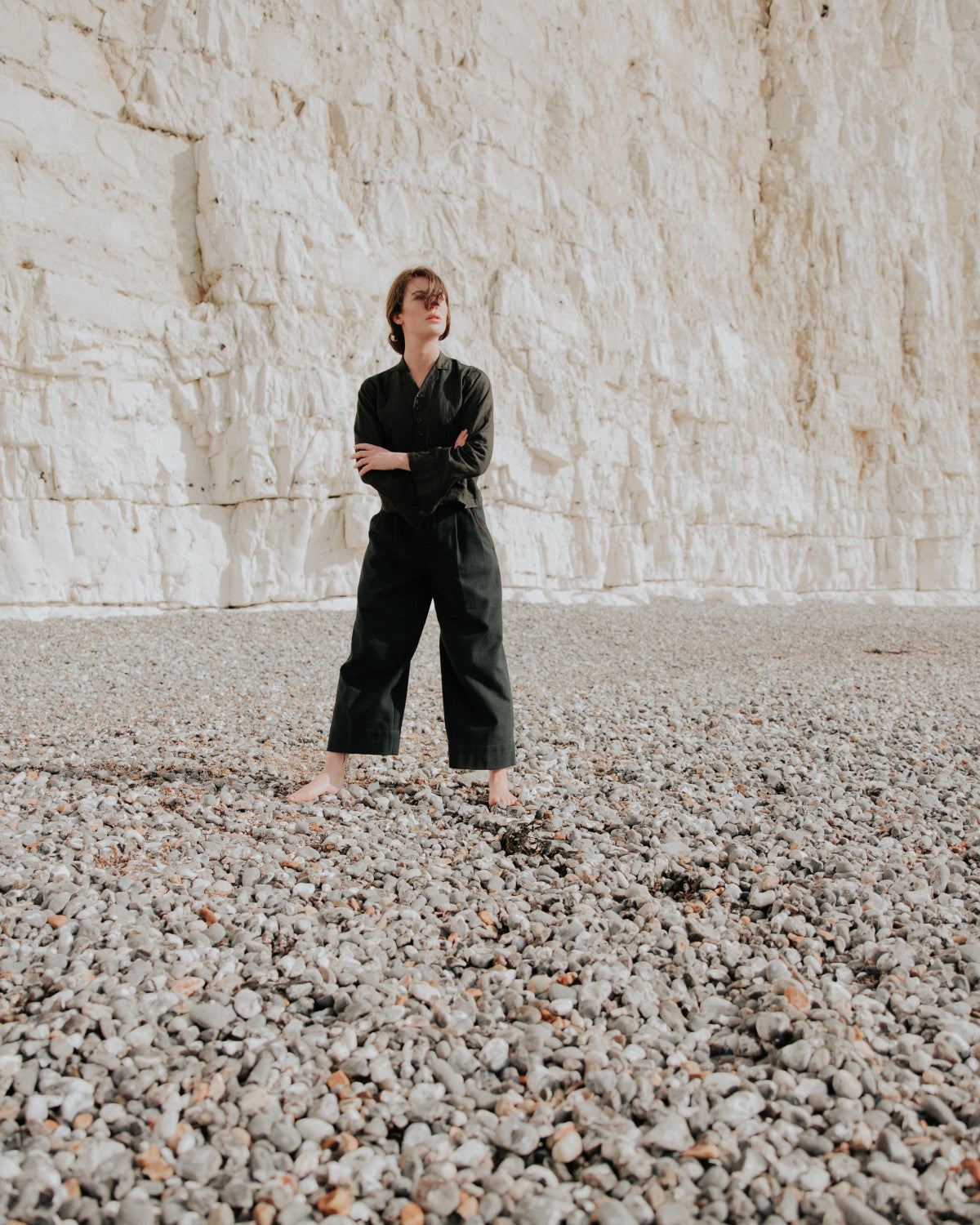 Person wearing a black outfit standing on a pebbly beach with a white cliff in the background