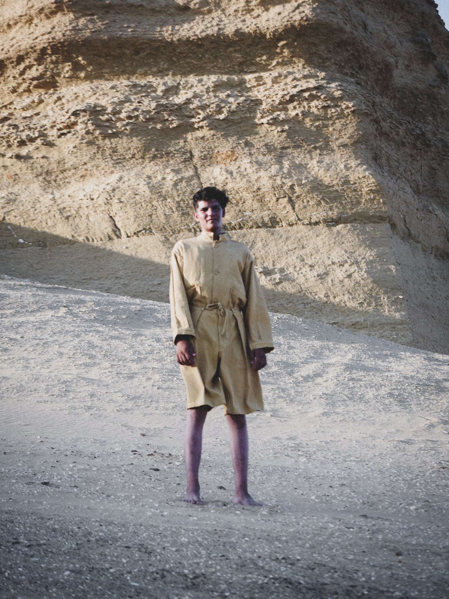 Person in a pale mustard suit standing in a desert landscape with a rocky cliff.