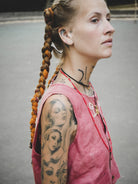 Woman with braided hair and tattoos wearing a dark pink vest on a plain background
