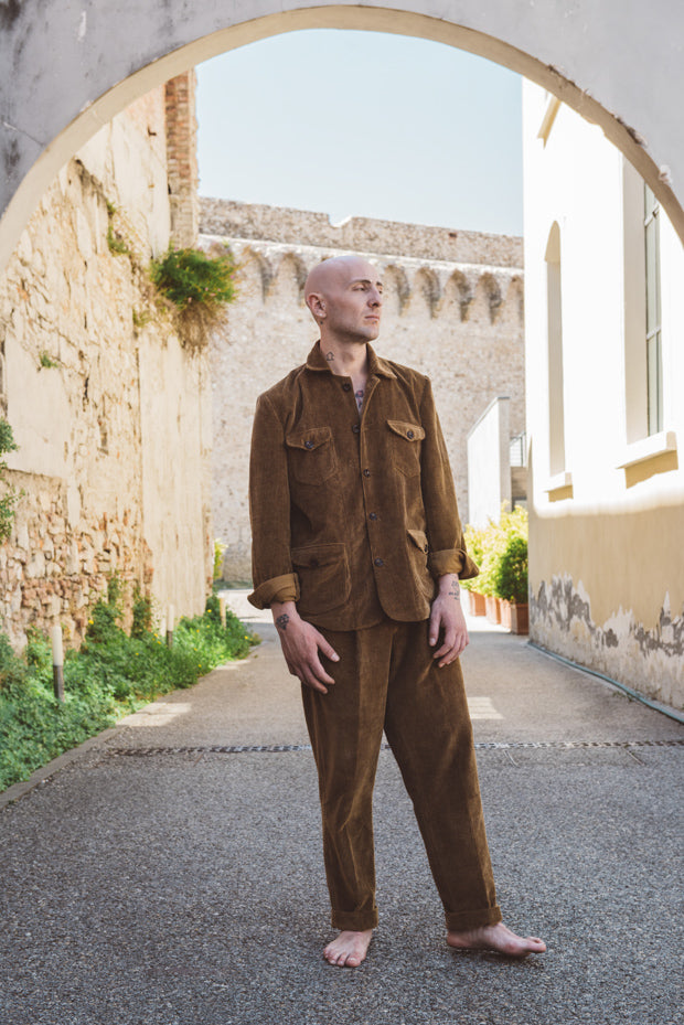 Man in a brown outfit standing in an alleyway with stone walls and plants.